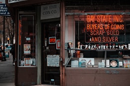 A storefront with various vintage and collectible items displayed. The signage includes a neon sign for a coin and precious metal buying establishment. There is an 'open' sign on the door, which is surrounded by various advertisements and posters. The window features a row of decorative figurines and collectibles.
