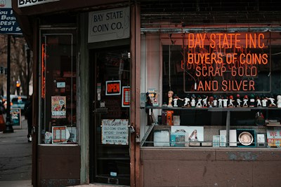 A storefront with various vintage and collectible items displayed. The signage includes a neon sign for a coin and precious metal buying establishment. There is an 'open' sign on the door, which is surrounded by various advertisements and posters. The window features a row of decorative figurines and collectibles.