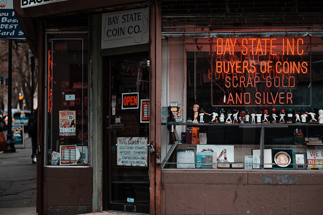 A storefront with various vintage and collectible items displayed. The signage includes a neon sign for a coin and precious metal buying establishment. There is an 'open' sign on the door, which is surrounded by various advertisements and posters. The window features a row of decorative figurines and collectibles.