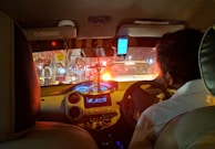 A nighttime scene inside a car seen from the back seat, with a person driving. The dashboard is illuminated, and a smartphone is mounted on the windshield. Red brake lights from cars ahead and reflections from city lights are visible through the windshield.
