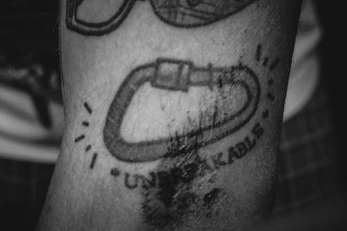 A black and white tattoo on a person's arm features a carabiner with the word 'UNBREAKABLE' underneath it. The tattoo is surrounded by small lines indicating movement or emphasis.