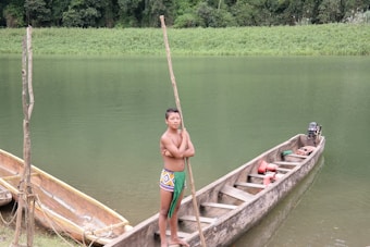 A young person stands in a wooden canoe by a calm, green river surrounded by lush vegetation. They hold a long pole and wear colorful, patterned clothing. Another canoe is moored nearby, and a small motor is visible at the back of the canoe they are in.