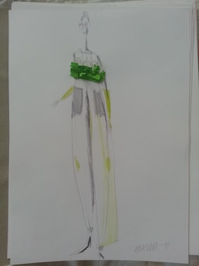 A fashion sketch showing a tall, slender figure wearing a flowing garment. The design features loose pants and an elaborate green embellishment around the waist area. The sketch is done in pencil with hints of green and yellow shading, emphasizing a modern and artistic style.