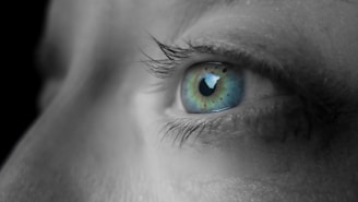 A close-up of a human eye with striking green and blue hues, the pupil is centered, and the surrounding skin appears soft and slightly out of focus.