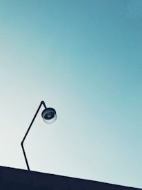 A minimalist photograph featuring a single street lamp rising from a slightly visible rooftop against a clear blue sky. The lamp has a modern, simplistic design with a curved arm. The image captures a sense of simplicity and openness.