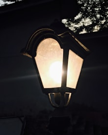 A vintage-style outdoor lantern with a bright light glowing inside, casting a soft glow. The background is dark with silhouettes of trees partially visible against the dim sky.