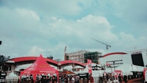 An outdoor scene at a public event with several tents, including a prominent red tent branded with smartmotor.co.id. A construction crane is visible alongside buildings in the background, one with 'PT Jakarta International Expo' signage. People are gathered around, indicating a busy atmosphere.