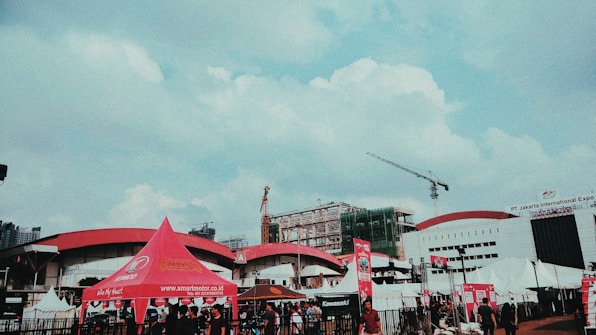 An outdoor scene at a public event with several tents, including a prominent red tent branded with smartmotor.co.id. A construction crane is visible alongside buildings in the background, one with 'PT Jakarta International Expo' signage. People are gathered around, indicating a busy atmosphere.