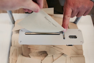 A person's hand is inserting an envelope into a metal slot on a box filled with similar envelopes. The envelopes appear to be related to voting or a formal procedure.
