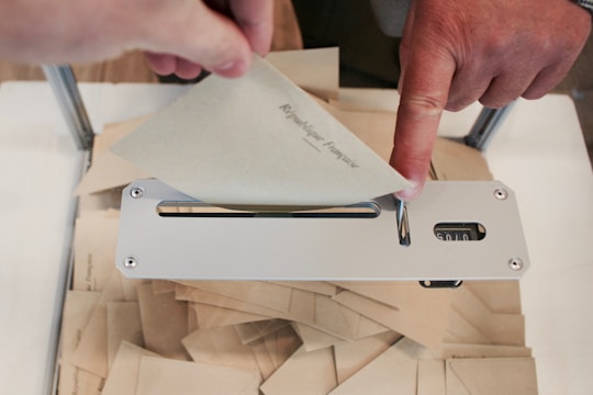 A person's hand is inserting an envelope into a metal slot on a box filled with similar envelopes. The envelopes appear to be related to voting or a formal procedure.