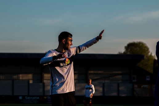A person in a white soccer jersey with black accents is captured mid-action on a soccer field during a game. The setting is outdoor, with a structure in the background and trees visible under a blue sky. The lighting is low, suggesting either early morning or late afternoon.