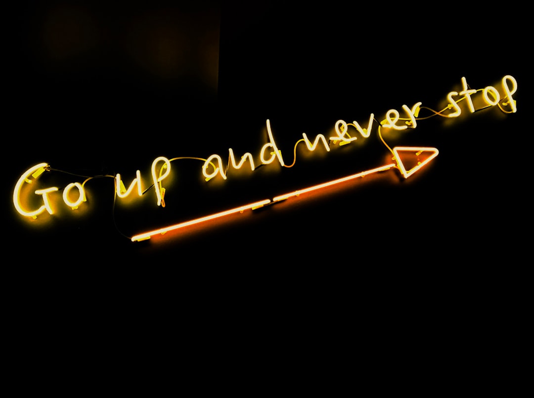 A yellow “Go up and never stop” neon with a long arrow against a black background