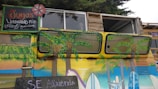 A vibrantly painted vehicle featuring tropical and beach-themed artwork, including palm trees and surfboards. Above the vehicle is a hand-painted sign advertising juices and mixed lemonades in Spanish. The sign has colorful lettering and features a small flower design. The van is parked in front of a rustic wooden structure with windows above.