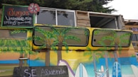 A vibrantly painted vehicle featuring tropical and beach-themed artwork, including palm trees and surfboards. Above the vehicle is a hand-painted sign advertising juices and mixed lemonades in Spanish. The sign has colorful lettering and features a small flower design. The van is parked in front of a rustic wooden structure with windows above.