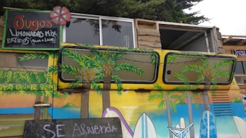 A vibrantly painted vehicle featuring tropical and beach-themed artwork, including palm trees and surfboards. Above the vehicle is a hand-painted sign advertising juices and mixed lemonades in Spanish. The sign has colorful lettering and features a small flower design. The van is parked in front of a rustic wooden structure with windows above.