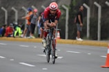 A cyclist wearing a red outfit and helmet is riding a bicycle on a road. The road is lined with cones, and spectators can be seen standing and sitting on the sidewalk in the background. The scene appears to be part of a cycling event.