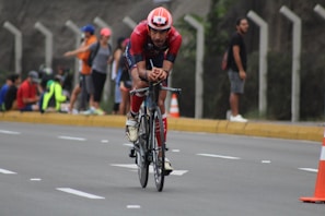A cyclist wearing a red outfit and helmet is riding a bicycle on a road. The road is lined with cones, and spectators can be seen standing and sitting on the sidewalk in the background. The scene appears to be part of a cycling event.