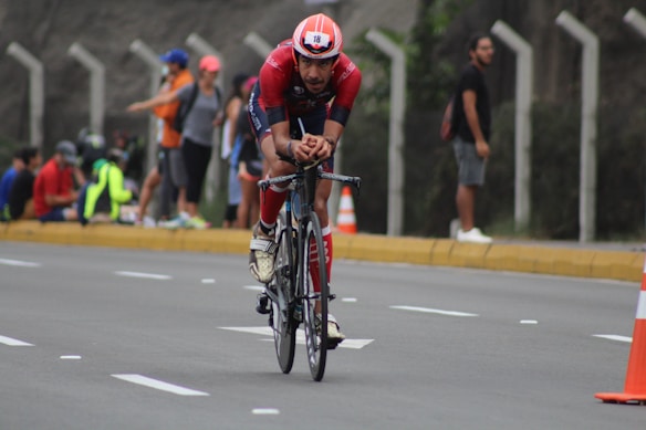 A cyclist wearing a red outfit and helmet is riding a bicycle on a road. The road is lined with cones, and spectators can be seen standing and sitting on the sidewalk in the background. The scene appears to be part of a cycling event.