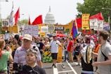 A large crowd of people are gathered in a protest or rally, holding various colorful signs and banners advocating for climate justice and environmental protection. Many individuals are wearing hats and sunglasses, and some are holding flags. In the background, the United States Capitol building is visible, suggesting the event is taking place in Washington D.C. The atmosphere is lively and passionate.