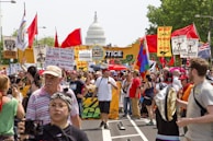 A large crowd of people are gathered in a protest or rally, holding various colorful signs and banners advocating for climate justice and environmental protection. Many individuals are wearing hats and sunglasses, and some are holding flags. In the background, the United States Capitol building is visible, suggesting the event is taking place in Washington D.C. The atmosphere is lively and passionate.