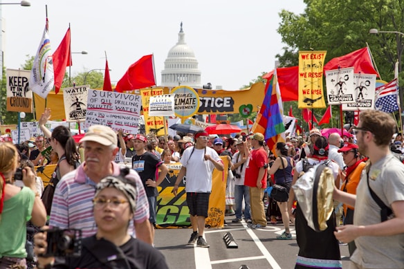 A large crowd of people are gathered in a protest or rally, holding various colorful signs and banners advocating for climate justice and environmental protection. Many individuals are wearing hats and sunglasses, and some are holding flags. In the background, the United States Capitol building is visible, suggesting the event is taking place in Washington D.C. The atmosphere is lively and passionate.