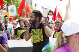 A group of people are participating in a protest, with many holding red flags and signs. The focal point is a man speaking passionately into a megaphone, wearing a shirt with '#BlackLivesMatter' printed on it. The scene is lively, with others in the group also wearing similar shirts, and there is a sense of collective action and unity.