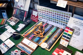 A display of art supplies including a wide variety of watercolor paints organized in a wooden box, colored pencils, and brushes. A color chart is prominently featured at the back, and a wooden mannequin rests on top of the paints. Additional items like waterbrushes and booklets are scattered around.