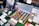 A display of art supplies including a wide variety of watercolor paints organized in a wooden box, colored pencils, and brushes. A color chart is prominently featured at the back, and a wooden mannequin rests on top of the paints. Additional items like waterbrushes and booklets are scattered around.