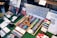 A display of art supplies including a wide variety of watercolor paints organized in a wooden box, colored pencils, and brushes. A color chart is prominently featured at the back, and a wooden mannequin rests on top of the paints. Additional items like waterbrushes and booklets are scattered around.