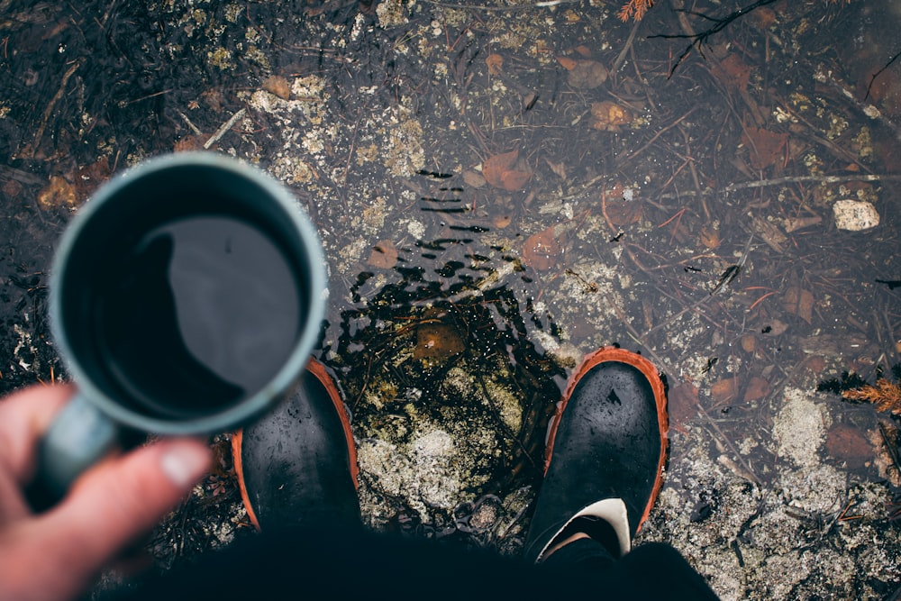 person holding coffee mug with pair of black shoes during daytime