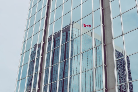 A reflection of a skyscraper in the large glass windows of another building, displaying a Canadian flag prominently in one of the panes. The setting suggests a modern urban environment with minimalistic architectural elements.