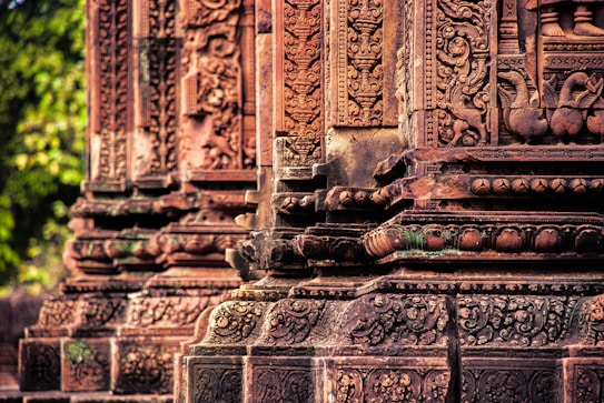 Intricate stone carvings on ancient temple structures, showcasing detailed patterns and figures in a reddish-brown hue. The carvings include decorative motifs and symbolic figures engraved in the stone pillars.