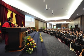 A large hall filled with people, where a graduation ceremony is taking place. A person in academic regalia is speaking at a podium on the left side, while graduates in caps and gowns are seated facing forward. The hall is decorated with flowers and has a large screen displaying an image of the speaker.