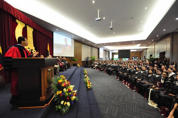 A large hall filled with people, where a graduation ceremony is taking place. A person in academic regalia is speaking at a podium on the left side, while graduates in caps and gowns are seated facing forward. The hall is decorated with flowers and has a large screen displaying an image of the speaker.
