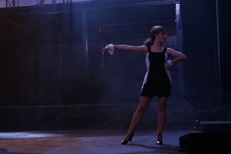 A woman in a black and white dress is striking a dance pose on a dimly lit stage. Her arm is extended gracefully, and she wears high heels, suggesting a theatrical or performance setting with dramatic lighting.