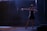 A woman in a black and white dress is striking a dance pose on a dimly lit stage. Her arm is extended gracefully, and she wears high heels, suggesting a theatrical or performance setting with dramatic lighting.