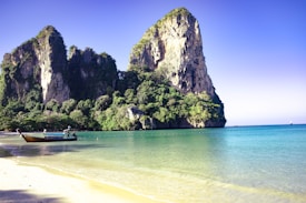 A picturesque coastal scene featuring a large limestone cliff covered in lush greenery, rising dramatically from the clear turquoise waters. A traditional wooden long-tail boat is seen floating on the gentle sea near the sandy beach, emphasizing the serene and tropical setting.