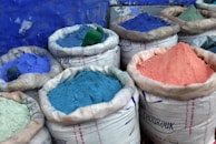 Several large sacks are filled with vibrant colored powders, including shades of blue, purple, and pink. The sacks are made of a white woven material and are set against a textured blue wall.