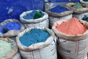 Several large sacks are filled with vibrant colored powders, including shades of blue, purple, and pink. The sacks are made of a white woven material and are set against a textured blue wall.