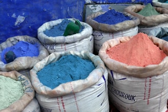 Several large sacks are filled with vibrant colored powders, including shades of blue, purple, and pink. The sacks are made of a white woven material and are set against a textured blue wall.