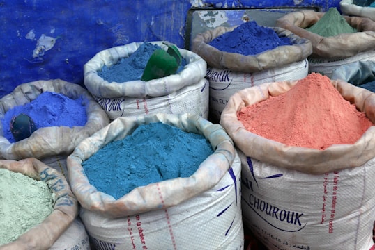 Several large sacks are filled with vibrant colored powders, including shades of blue, purple, and pink. The sacks are made of a white woven material and are set against a textured blue wall.