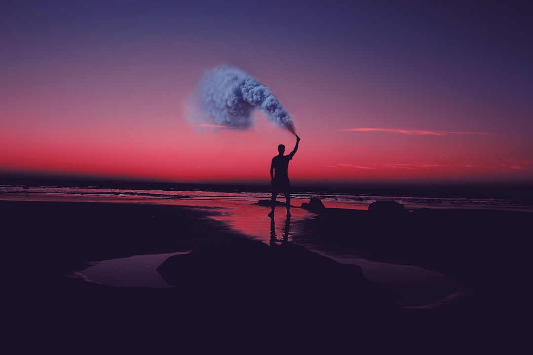 silhouette of man standing on seashore holding smoke can