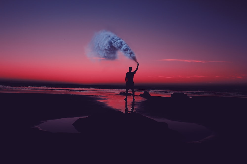 silhouette of man standing on seashore holding smoke can