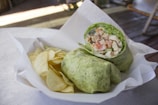 A green wrap filled with diced chicken, lettuce, and tomatoes is served on a white paper-lined tray alongside a handful of potato chips. The setting appears casual, with dim lighting and a wooden surface in the background, suggesting a relaxed dining atmosphere.