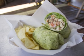 A green wrap filled with diced chicken, lettuce, and tomatoes is served on a white paper-lined tray alongside a handful of potato chips. The setting appears casual, with dim lighting and a wooden surface in the background, suggesting a relaxed dining atmosphere.