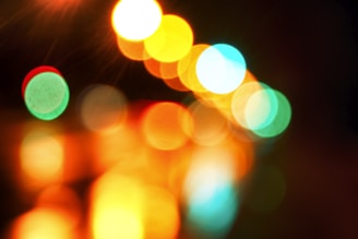 A photograph featuring abstract, out-of-focus, multi-colored lights. The light dots are of various sizes, primarily in shades of green, yellow, orange, red, and blue against a dark background. The image has a bokeh effect, creating a soft, dreamy atmosphere.