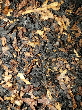 A close-up view of a mixture of dark and light brown dried leaves, possibly tobacco or a similar organic material. The leaves are finely shredded and layered on top of each other, creating a textured and chaotic arrangement.