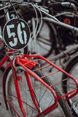 A vintage bicycle with a prominent red color is equipped with a round black number plate displaying the number 56. The focus is on the front part of the bicycle, including the handlebar and wheel. The background shows parts of other bicycles, creating a sense of a crowded bicycle stand.