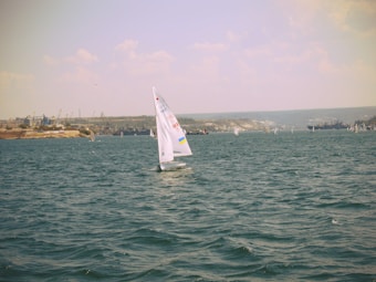 A sailboat is gliding through a vast expanse of water, with its sails catching the wind. Several other sailboats are visible in the distance, scattered across the water. The shoreline is lined with industrial structures, possibly cranes and storage tanks, and the horizon is misty under a partly cloudy sky.