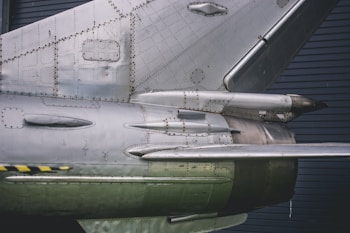 Close-up view of an aircraft's metallic structure, showcasing intricate details of the rivets and panels. The surface appears weathered, with a combination of matte and shiny metal finishes.